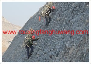 两个施工人员背着钢丝绳网在已经安装了的主动防护网的山坡上往上爬